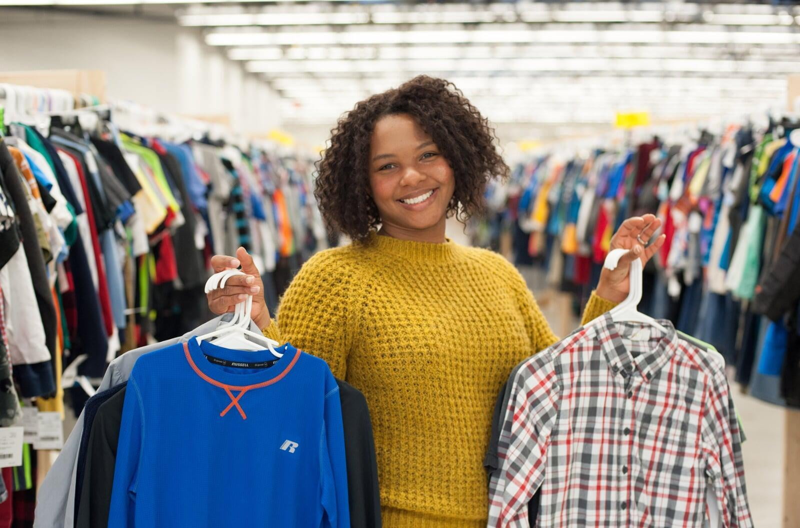 Mom wears baby and holds toddler boy clothes in each hand. Second mom shops behind her from the racks of boys clothes.